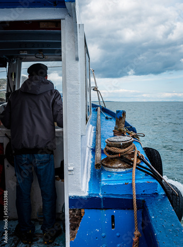Small fishing boat being sailed by an old fisherman visible from behind. Half view of cabin or cockpit and ocean on a fishing trawler shows the hardships of fishermen at sea with rusty boat equipment