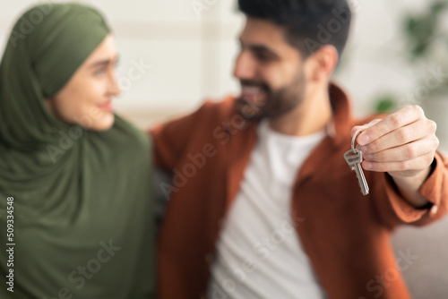 Happy Muslim Spouses Showing New Home Key Embracing Sitting Indoor