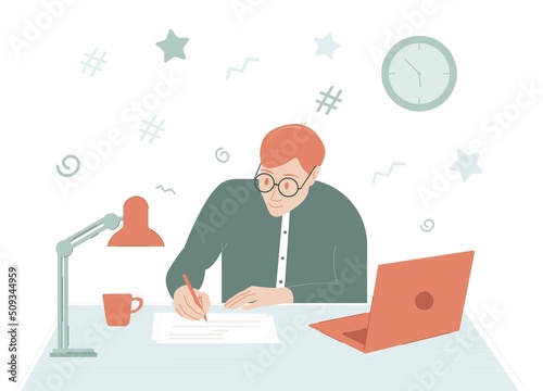 The student sits to study at the table. There is a lamp, a mug and a laptop on the table. A student writes something on a piece of paper with a pen