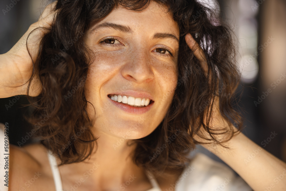 Close-up of happy young caucasian girl smiling looking into camera touching dark short hair. Brunette with freckles poses on blurry background. Concept of real emotion Close-up of happy young caucasian girl smiling looking into camera touching dark short hair. Brunette with freckles poses on blurry background. Concept of real emotion