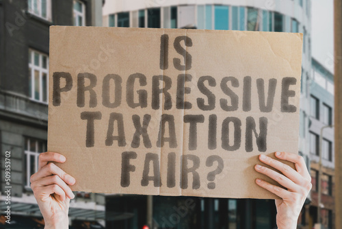 The question " Is progressive taxation fair? " is on a banner in men's hands with blurred background. Federal. Increase. Growing. Grow. Potential. Up. System. Trend. Return. Revenue. Sale. Office