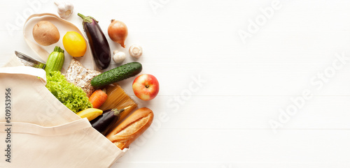 Cotton grocery tote bag with fresh vegetables, fruits, baguette and canned goods on white wooden background. Healthy food shopping, eco-friendly concept. Flat lay, copy space, banner.