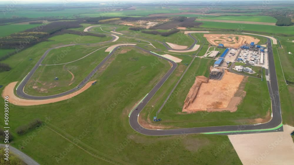 AERIAL: View of racing circuit and facilities with sport race cars training laps. Racing cars moving in clockwise direction of tarmac racecourse ring. Adrenaline motor sport for high-speed enthusiasts AERIAL: View of racing circuit and facilities with sport race cars training laps. Racing cars moving in clockwise direction of tarmac racecourse ring. Adrenaline motor sport for high-speed enthusiasts