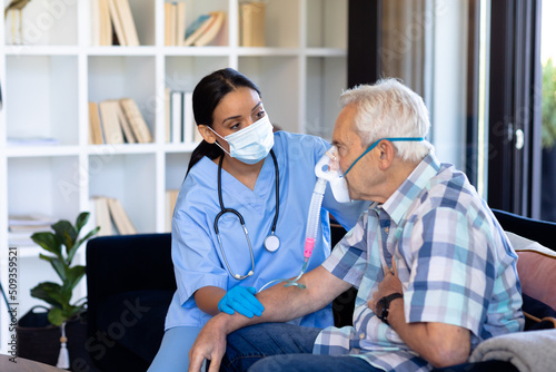 Biracial female doctor examining caucasian senior man wearing oxygen mask sitting on sofa at home