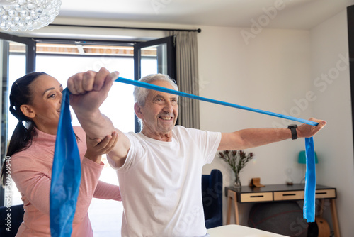 Biracial female physiotherapist assisting caucasian senior man stretching resistance band at home