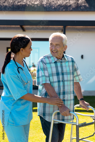 Biracial female doctor helping caucasian senior man in walking with walker in yard on sunny day