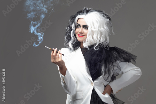 A fatal beauty in a daring fashion image with black and white hair. A rebellious stylish image for Halloween. a young woman in a black and white outfit smokes a cigarette using a mouthpiece