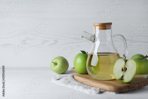 Jug of tasty juice and fresh ripe green apples on white wooden table, space for text photo