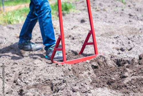 A farmer in jeans digs the ground with a red fork-shaped shovel. A miracle shovel, a handy tool. Manual cultivator. The cultivator is an efficient hand tool for tillage. Loosening the bed.