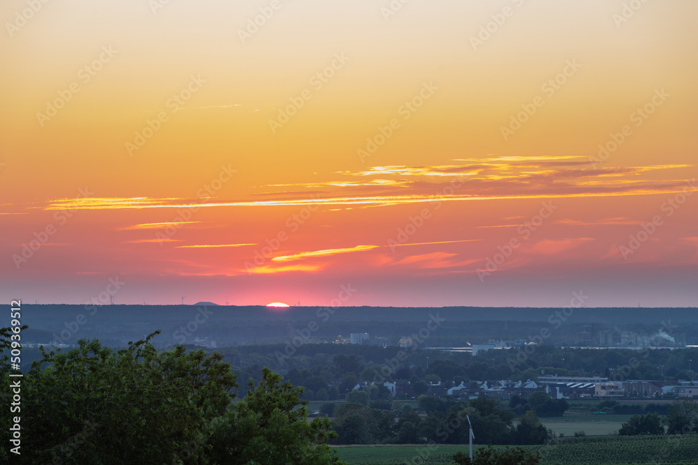 A wonderful sunset with a few fluffy clouds, covering the rolling hills of south Limburg in the Netherlands with an orange and golden glow, creating a magical atmosphere over the agricultural fields.