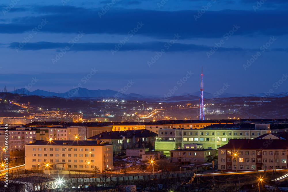 Evening city landscape. Buildings and TV tower view. Mountains in the distance. Evening twilight. Magadan, Magadan Region, Siberia, Russia.