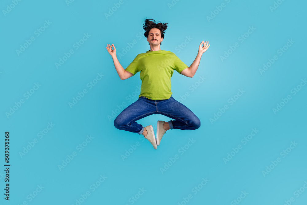 Full length photo of dreamy attractive man wear green t-shirt practicing yoga jumping high isolated blue color background Full length photo of dreamy attractive man wear green t-shirt practicing yoga jumping high isolated blue color background