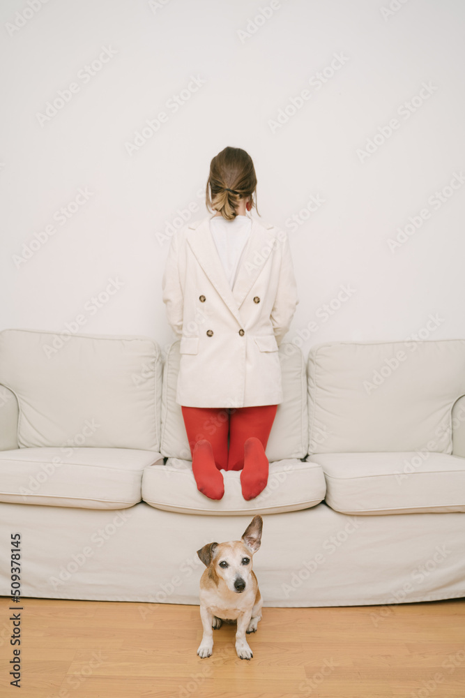 young girl in red tights and white jacket put on opposite is sitting on a beige sofa. a small dog sits on the floor and looks into the chamber. emotional state metaphor. Feeling lost and fake