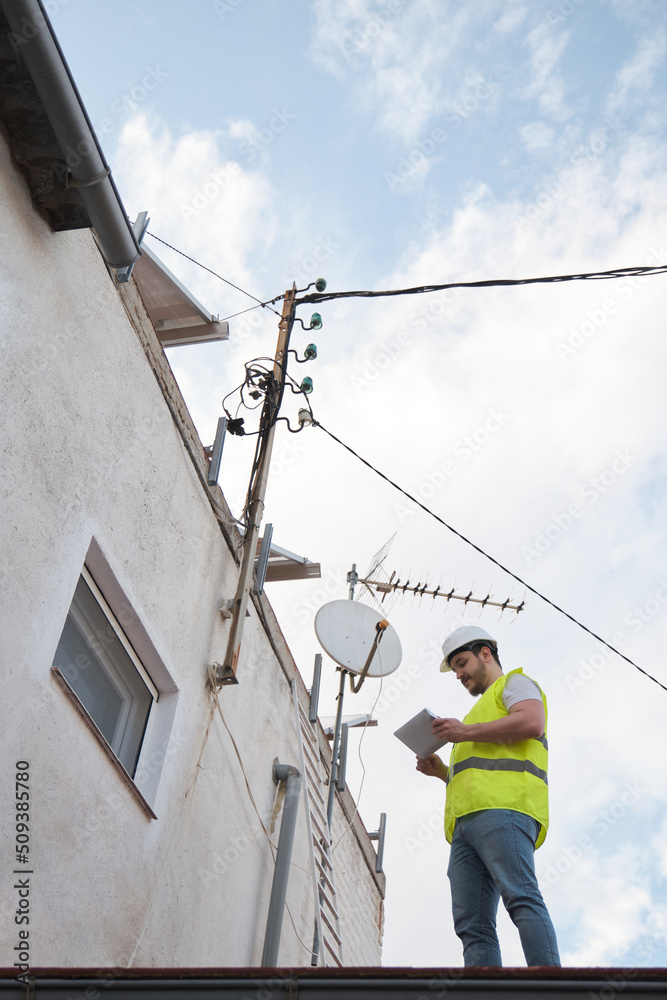 Technician checking electric wire power lines and telephone pole in a house roof of a residential area.