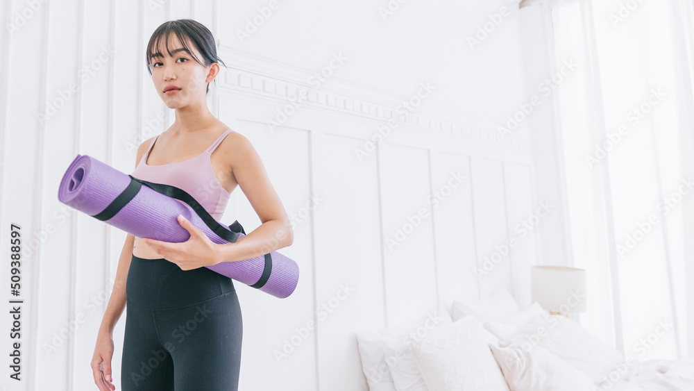Asian woman wearing sportswear and yoga pants carrying a rolled-up yoga mat standing in front of white wall background with sunlight from the window. Half-length image with copy space.