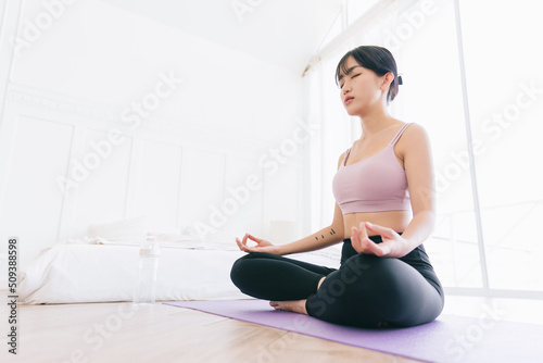 Young attractive sporty Asian woman practicing yoga on a yoga mat, doing Ardha Padmasana exercise, meditating in Half Lotus pose, indoor working out at home, wearing sportswear.