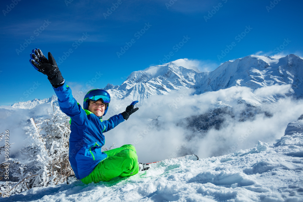 Smiling boy with alpine ski and helmet sit in snow lifting hands Smiling boy with alpine ski and helmet sit in snow lifting hands