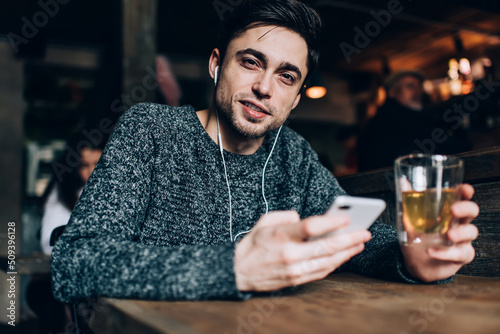 Portrait of young Caucaisan customer with beverage and smartphone in hands listening audio book during weekend daytime in cafe interior  millennial blogger in electronic headphones looking at camera