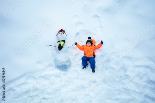 Boy in sport winter orange coat lay holding hand of snowman
