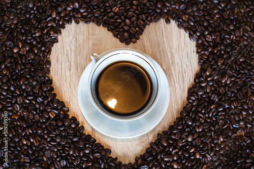 Fototapeta Naklejka Na Ścianę i Meble -  A cup of hot aromatic coffee in a saucer, against the background of scattered coffee beans in the form of a heart, on a wooden light brown table.