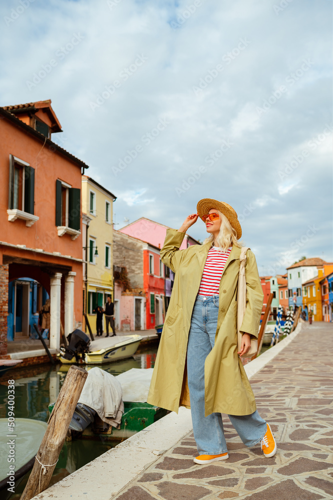 Happy smiling fashionable traveler woman walking among colorful houses on Burano island, Italy. Model wearing straw hat, trench coat, wide leg jeans, striped t-shirt, orange sneakers, sunglasses