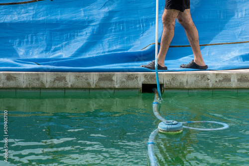 Piernas del hombre que hace mantenimiento en la piscina, lona recién quitada, agua verde, robot y flotador de cloro