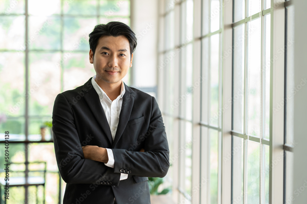 Portrait of Asian business man wear suit at the workspace office. Smiling Asian business man standing with arms crossed in background modern office Portrait of Asian business man wear suit at the workspace office. Smiling Asian business man standing with arms crossed in background modern office