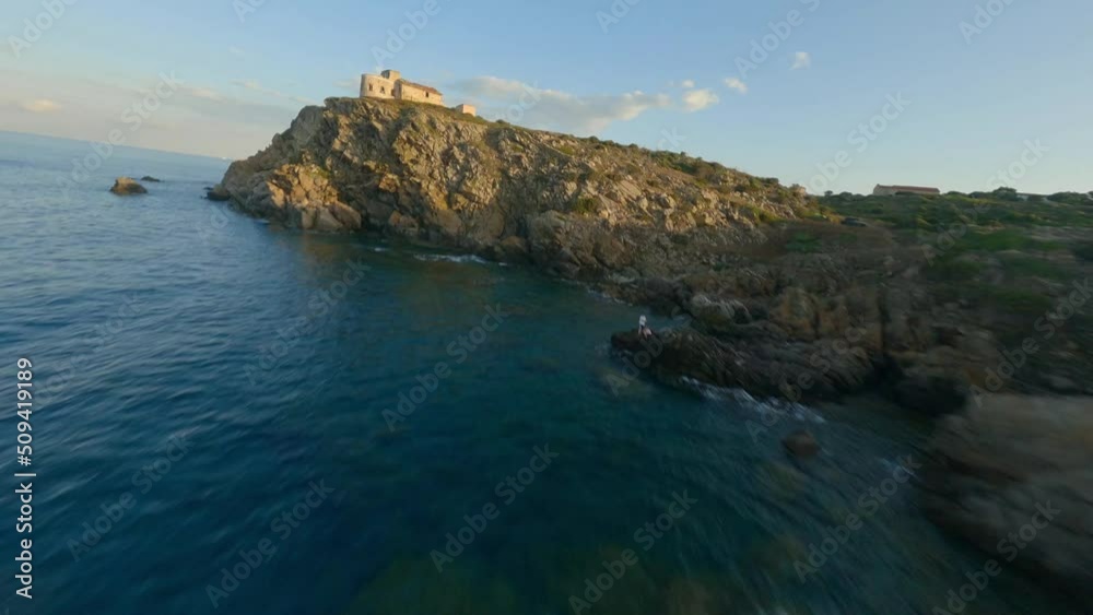 FPV video, view from above, stunning aerial view from an FPV drone flying at high speed over a rocky coastline with a lighthouse illuminated during a dramatic sunset. Faro di Capo Ferro, Sardinia.