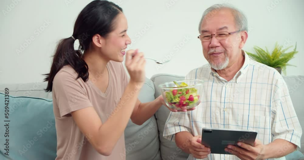 Caregiver young Asian woman feeding vegetable elderly Asian man sitting on sofa in living room at home.Woman therapist are monitoring the symptoms of senior male patient.Healthcare medical concept. Caregiver young Asian woman feeding vegetable elderly Asian man sitting on sofa in living room at home.Woman therapist are monitoring the symptoms of senior male patient.Healthcare medical concept.