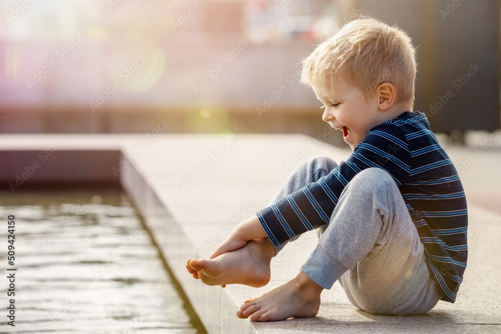 A curious child, a boy, barefoot touches the water of a city fountain. Hot and sunny summer day, the water is a good refreshment for the boy's feet. A curious child, a boy, barefoot touches the water of a city fountain. Hot and sunny summer day, the water is a good refreshment for the boy's feet.