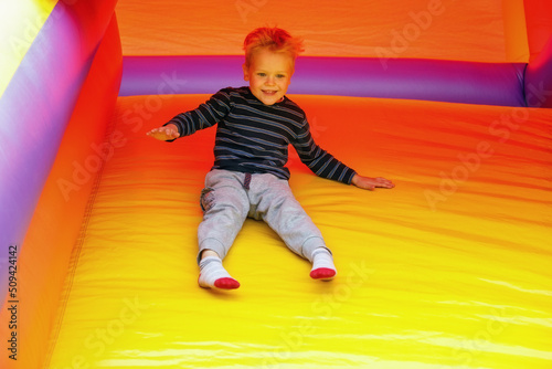 A cheerful, laughing boy sitting and slide down on a bright yellow rubber trampoline. Image in motion, a brave child is not afraid of anything