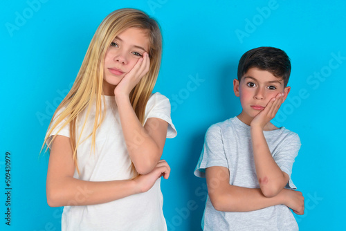 Very bored two kids boy and girl standing over blue studio background holding hand on cheek while support it with another crossed hand, looking tired and sick.