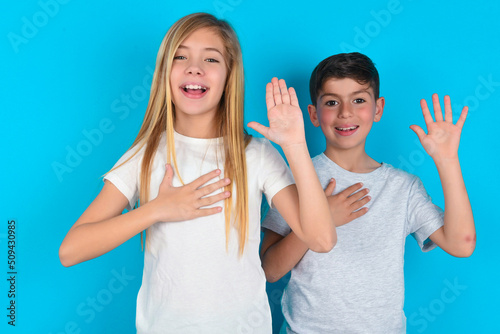 I swear, promise you not regret. Portrait of sincere two kids boy and girl standing over blue studio background raising one arm and hold hand on heart as give oath, telling truth, want you to believe.