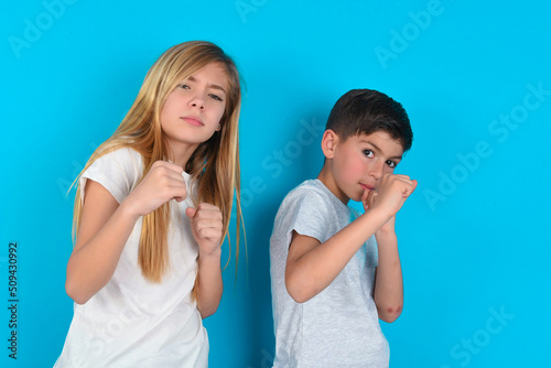 Portrait of attractive two kids boy and girl standing over blue studio background holding hands in front of him in boxing position going to fight.