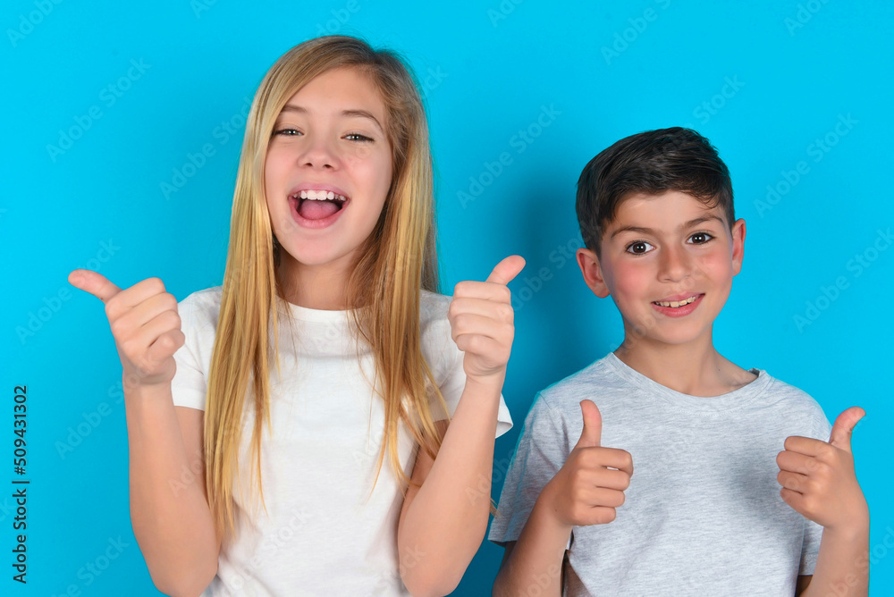 Optimistic two kids boy and girl standing over blue studio background showing thumbs up with positive emotions. Quality and recommendation concept. Optimistic two kids boy and girl standing over blue studio background showing thumbs up with positive emotions. Quality and recommendation concept.