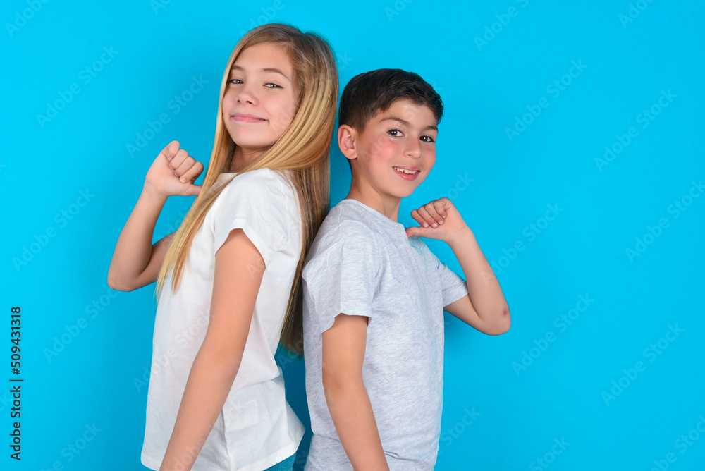 Closeup of cheerful two kids boy and girl standing over blue studio background looks joyful, satisfied and confident, points at himself with thumb. Closeup of cheerful two kids boy and girl standing over blue studio background looks joyful, satisfied and confident, points at himself with thumb.