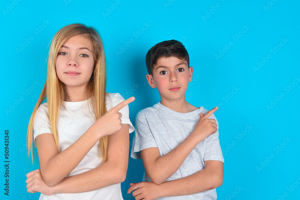 two kids boy and girl standing over blue studio background smiling broadly at camera, pointing fingers away, showing something interesting and exciting.