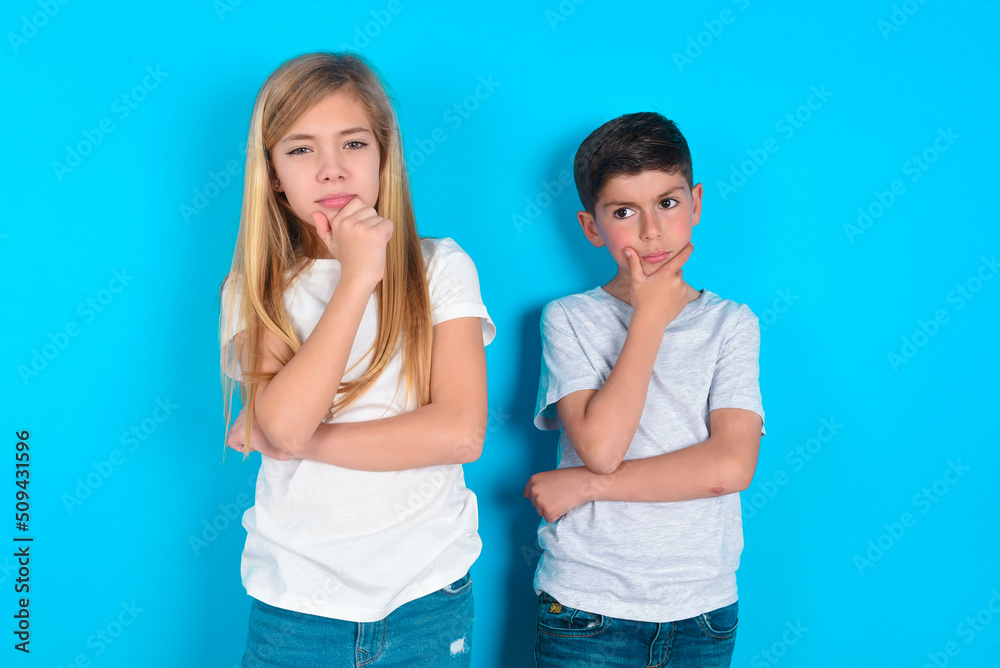 Thoughtful smiling two kids boy and girl standing over blue studio background keeps hand under chin, looks directly at camera, listens something with interest. Youth concept. Thoughtful smiling two kids boy and girl standing over blue studio background keeps hand under chin, looks directly at camera, listens something with interest. Youth concept.