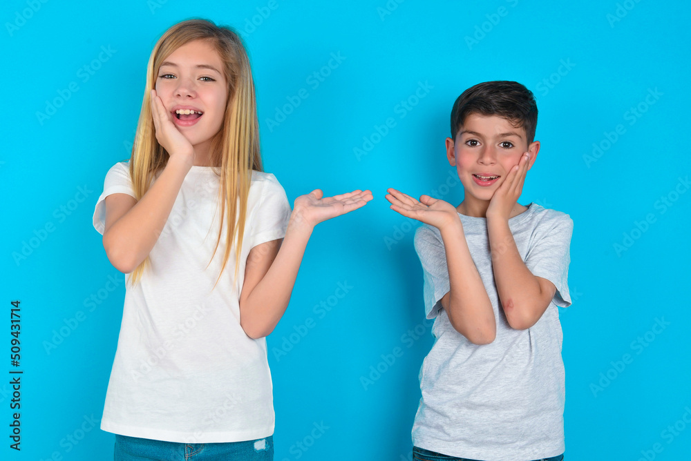 Positive glad two kids boy and girl standing over blue studio background says: wow how exciting it is, indicates something. One hand on his head and pointing with other hand. Positive glad two kids boy and girl standing over blue studio background says: wow how exciting it is, indicates something. One hand on his head and pointing with other hand.