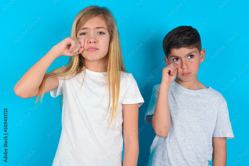 Unhappy two kids boy and girl standing over blue studio background crying while posing at camera whipping tears with hand. Unhappy two kids boy and girl standing over blue studio background crying while posing at camera whipping tears with hand.