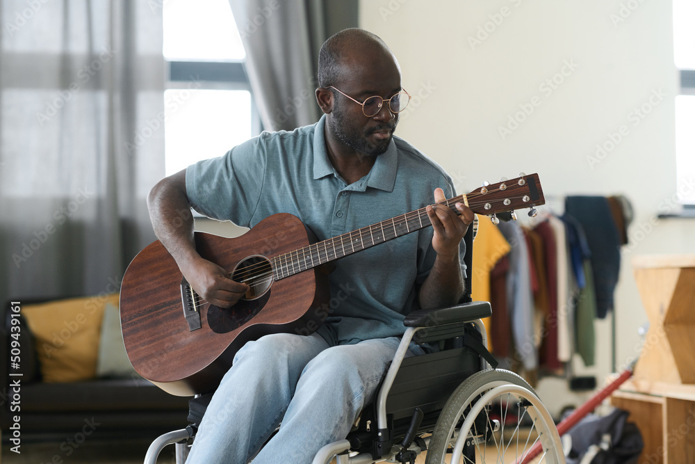 Fototapeta premium African American man in eyeglasses sitting in wheelchair and learning to play guitar during his leisure time at home