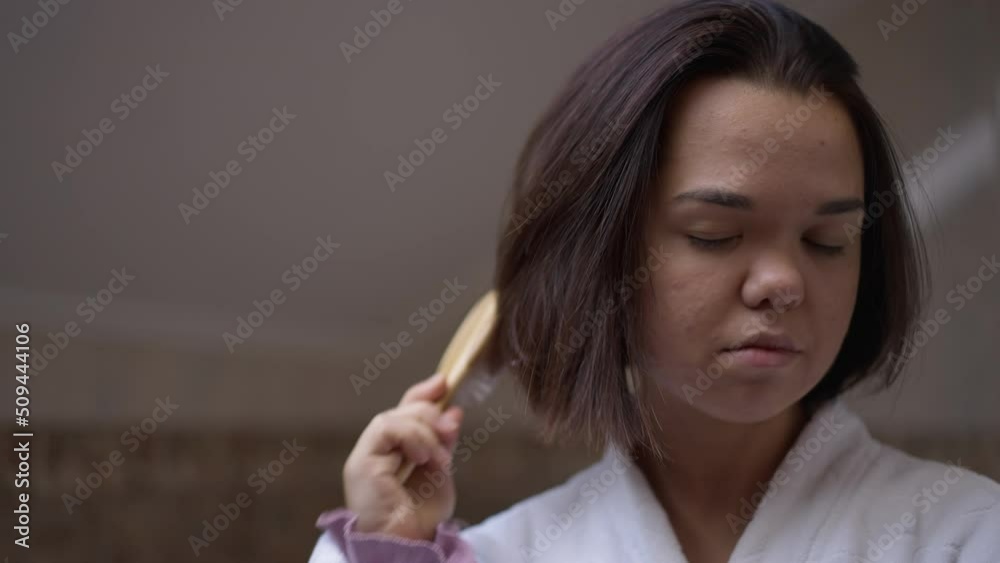 Close-up portrait of young little woman combing hair standing in bathroom at home. Concentrated Caucasian little person taking care of hairstyle indoors in the morning Close-up portrait of young little woman combing hair standing in bathroom at home. Concentrated Caucasian little person taking care of hairstyle indoors in the morning