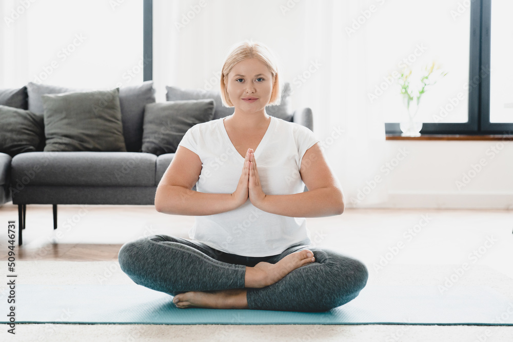 Zen-like caucasian young plus size plump woman in fitness clothes meditating on sporty mat, sitting relaxing in yoga lotus position at home looking at camera Zen-like caucasian young plus size plump woman in fitness clothes meditating on sporty mat, sitting relaxing in yoga lotus position at home looking at camera