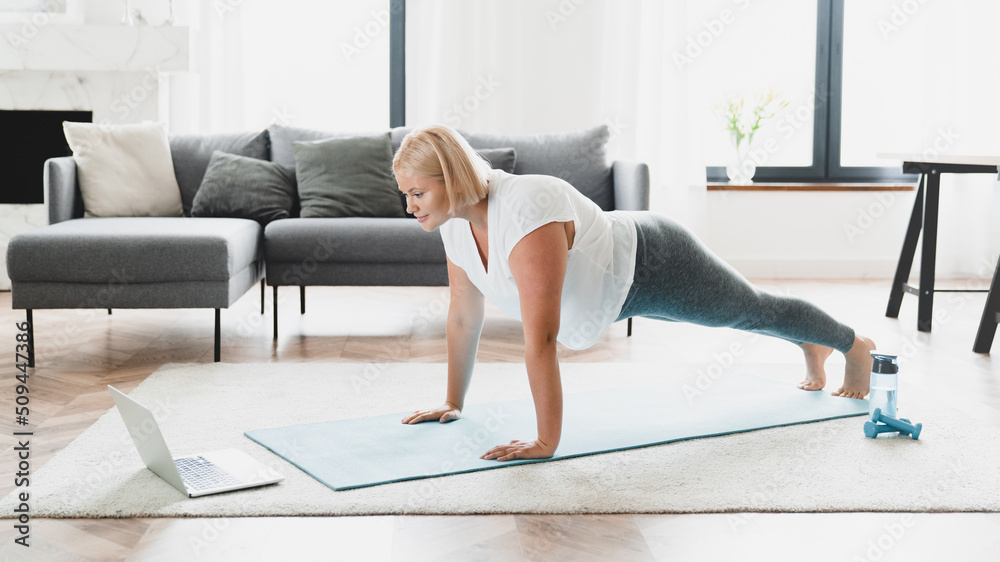 Young caucasian plus size plump woman athlete in fitness clothes standing in plank position, training stamina, slimming, burning calories watching online video tutorial at home Young caucasian plus size plump woman athlete in fitness clothes standing in plank position, training stamina, slimming, burning calories watching online video tutorial at home