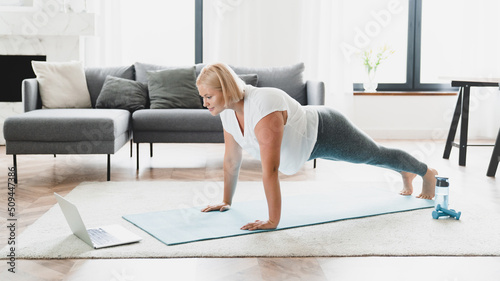 Young caucasian plus size plump woman athlete in fitness clothes standing in plank position, training stamina, slimming, burning calories watching online video tutorial at home