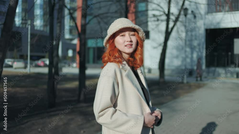A young woman of Asian appearance walks in the city park. She is wearing a light coat and a hat. A red-haired beautiful lady. Slow motion. A young woman of Asian appearance walks in the city park. She is wearing a light coat and a hat. A red-haired beautiful lady. Slow motion.