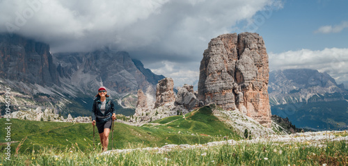 Canvas-taulu Smiling female trekker walking with backpack and trekking poles by green mountain hill with picturesque Dolomite Alps Cinque Torri formation on the background