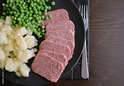 Sliced canned corned beef with boiled potatoes and peas on a black plate. photo