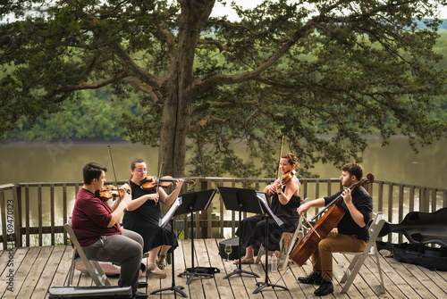 String quartet of two young men and two young women playing concert on wooden deck above Missouri River on summer evening; river and woods in background