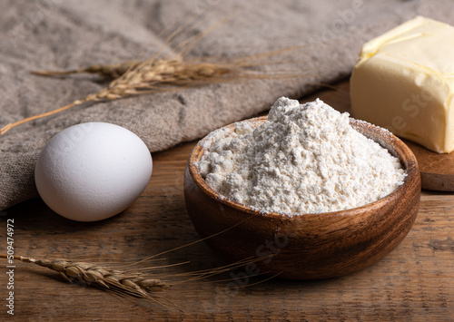Flour in a wooden bowl, butter and an egg on a wooden table.